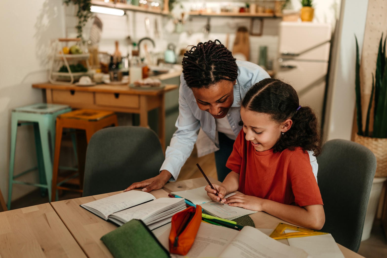 Mãe acompanhando a filha nos estudos em casa, à mesa com cadernos e livros